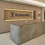 Graphic printed behind a reception desk in a building lobby that reads Rothschild in black text. 