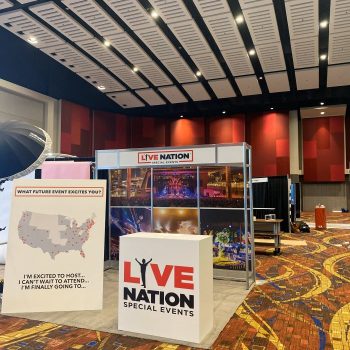 Live Nation Special Events trade show booth inside a convention hall, featuring branded signage, photo panels of concerts, and a white display counter on a patterned carpet floor.
