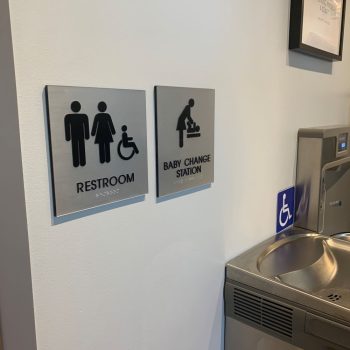 Brushed metal ADA restroom and baby change station signs mounted on white wall in Chicago Downtown.