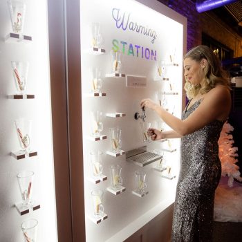 Woman using a custom illuminated warming station display with glass mugs mounted on a white backlit wall at an indoor event.