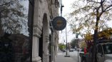 Round blade sign mounted on a historic stone storefront along a city street with trees and traffic.
