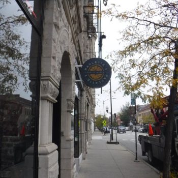 Round blade sign mounted on a historic stone storefront along a city street with trees and traffic.