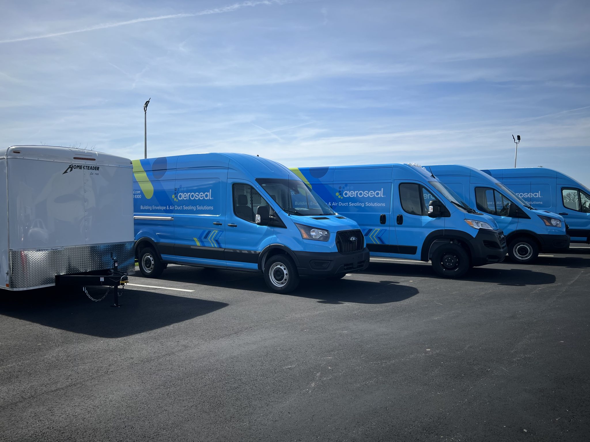 Three blue commercial fleet vans with full vehicle wraps parked beside a white trailer in Dayton,