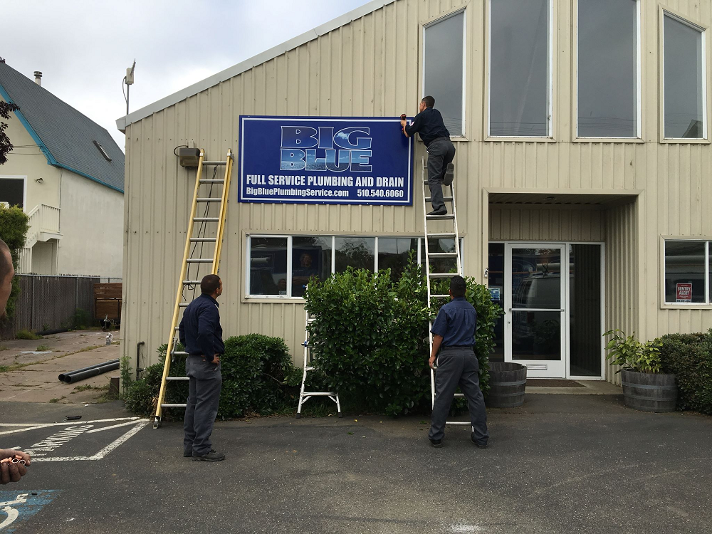 Workers hanging a business sign for Big Blue plumbing outside of a building.