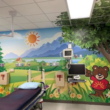 Children's hospital exam room with a mural of a cartoon meadow scene and the tree extending to the ceiling tiles.