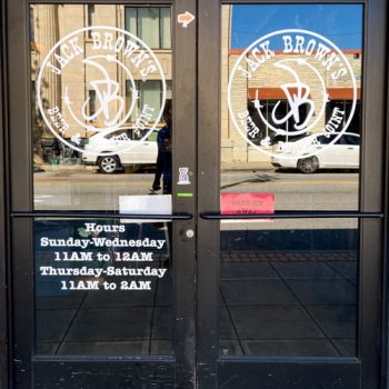 Storefront logos and restaurant hours in white vinyl on Jack Brown's front doors in downtown Greenville, SC