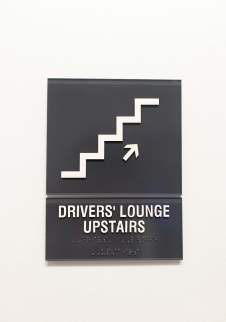 Dark grey custom ADA sign with white text and graphics directing to the driver's lounge.