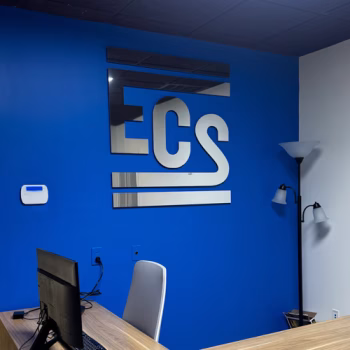 Polished aluminum dimensional logo installed on a painted blue wall in ECS's lobby.