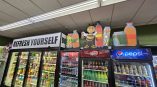 Custom die-cut and shaped retail graphics featuring beverage bottles and a cartoon bee mascot, installed on top of commercial refrigerators for branding, a project by SpeedPro Irving.
