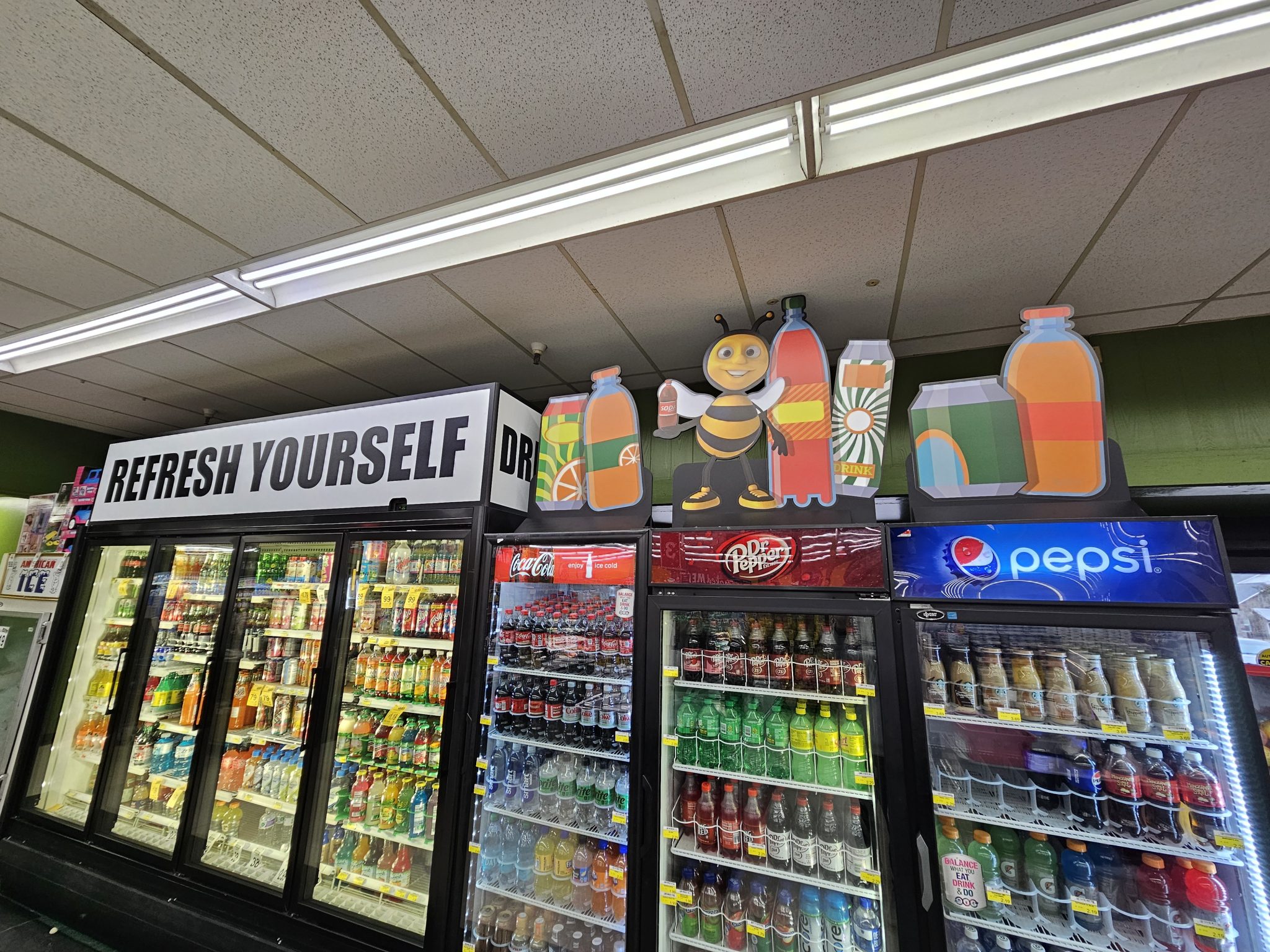 Custom die-cut and shaped retail graphics featuring beverage bottles and a cartoon bee mascot, installed on top of commercial refrigerators for branding, a project by SpeedPro Irving.