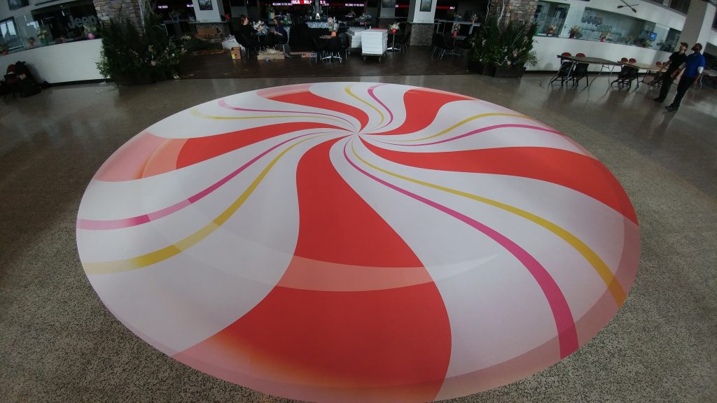 Large circular floor graphic with red, white, pink, and yellow swirling stripes resembling a peppermint candy in an open indoor space.