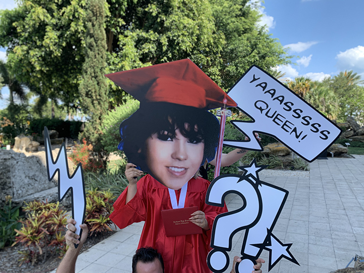 young woman wearing red graduation uniform holding up cutout sign of a woman's face
