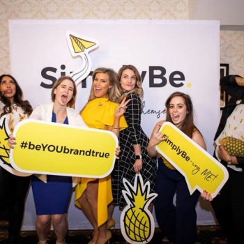 6 women posing in front of a SimplyBe branded backdrop while holding SimpleBe branded decorations