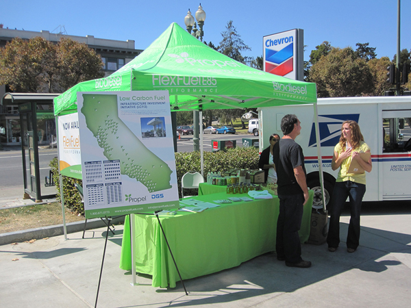 Green tent, poster and table set up for FlexFuelE85 in a Chevron parking lot with a woman and man standing in front.