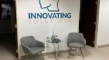 Two gray chairs and a glass table in front of a white wall with an outline of a head that says “innovating solutions”.