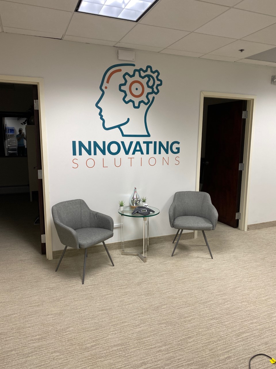 Two gray chairs and a glass table in front of a white wall with an outline of a head that says “innovating solutions”.
