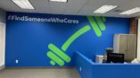 Bright blue wall that says “#FindSomeoneWhoCares” in white with green dumbbell on it next to a desk. 