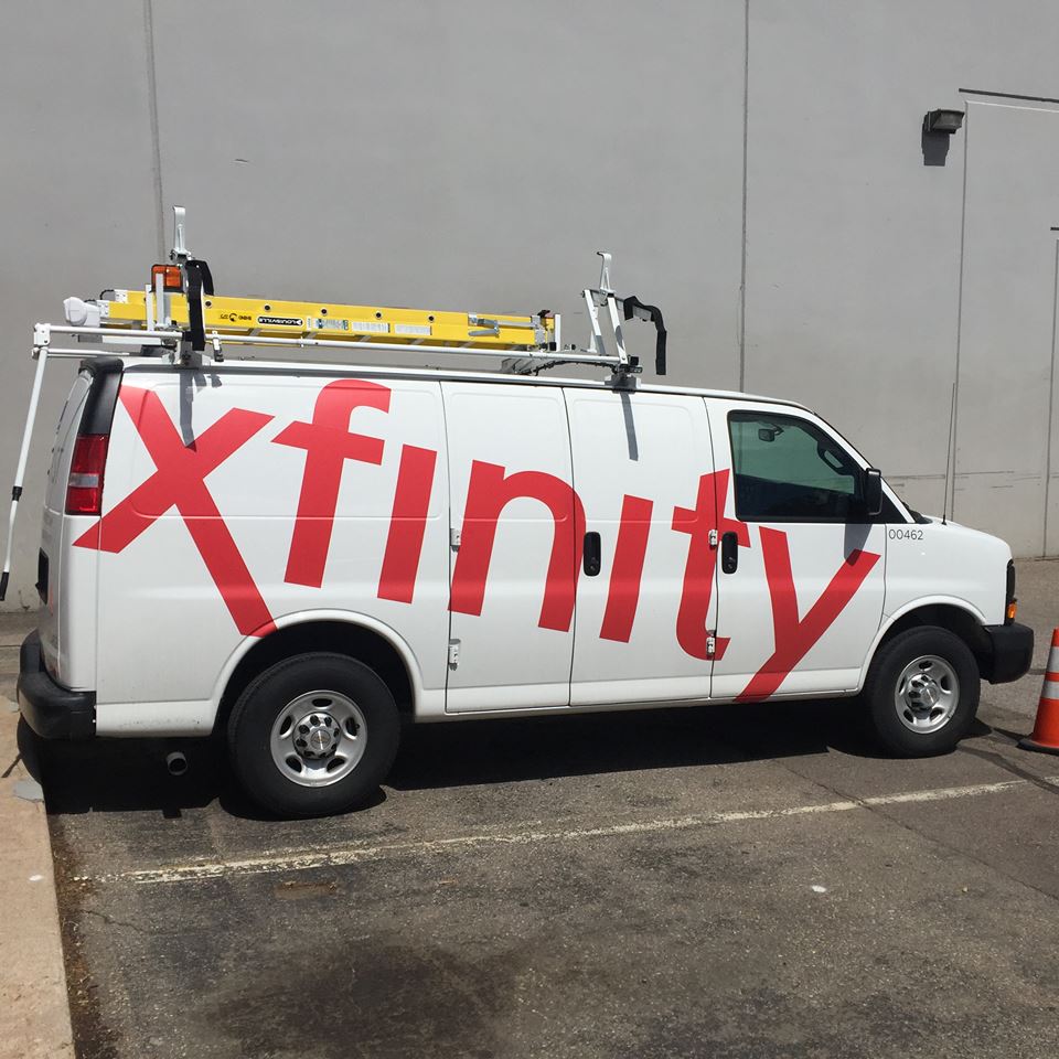Yellow ladder on top of a white van with red decal that says Xfinity going across the doors.