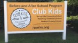 Bright orange yard sign promoting the 'Club Kids Before and After School Program' with school names and website.