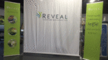 Trade show backdrop displaying the REVEAL brand logo and tagline, flanked by retractable banners with methodology details.