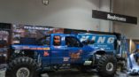 Full custom monster truck exhibit with oversized tires and vibrant blue graphics featuring a cartoon character at a trade show.