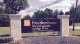 Monument sign for Freedom House displaying service times and website, installed on brick structure with landscaped surroundings.