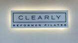 Dimensional acrylic lobby sign for Clearly Reformer Pilates, illuminated with backlighting on a white wall.