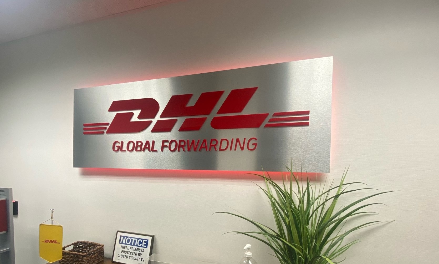 Dimensional acrylic sign featuring the DHL logo and 'GLOBAL FORWARDING' text with illuminated edges in an office lobby.