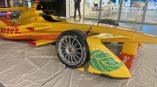 DHL-branded race car displayed on a white mat, featuring yellow and red graphics and leaf design on the side.