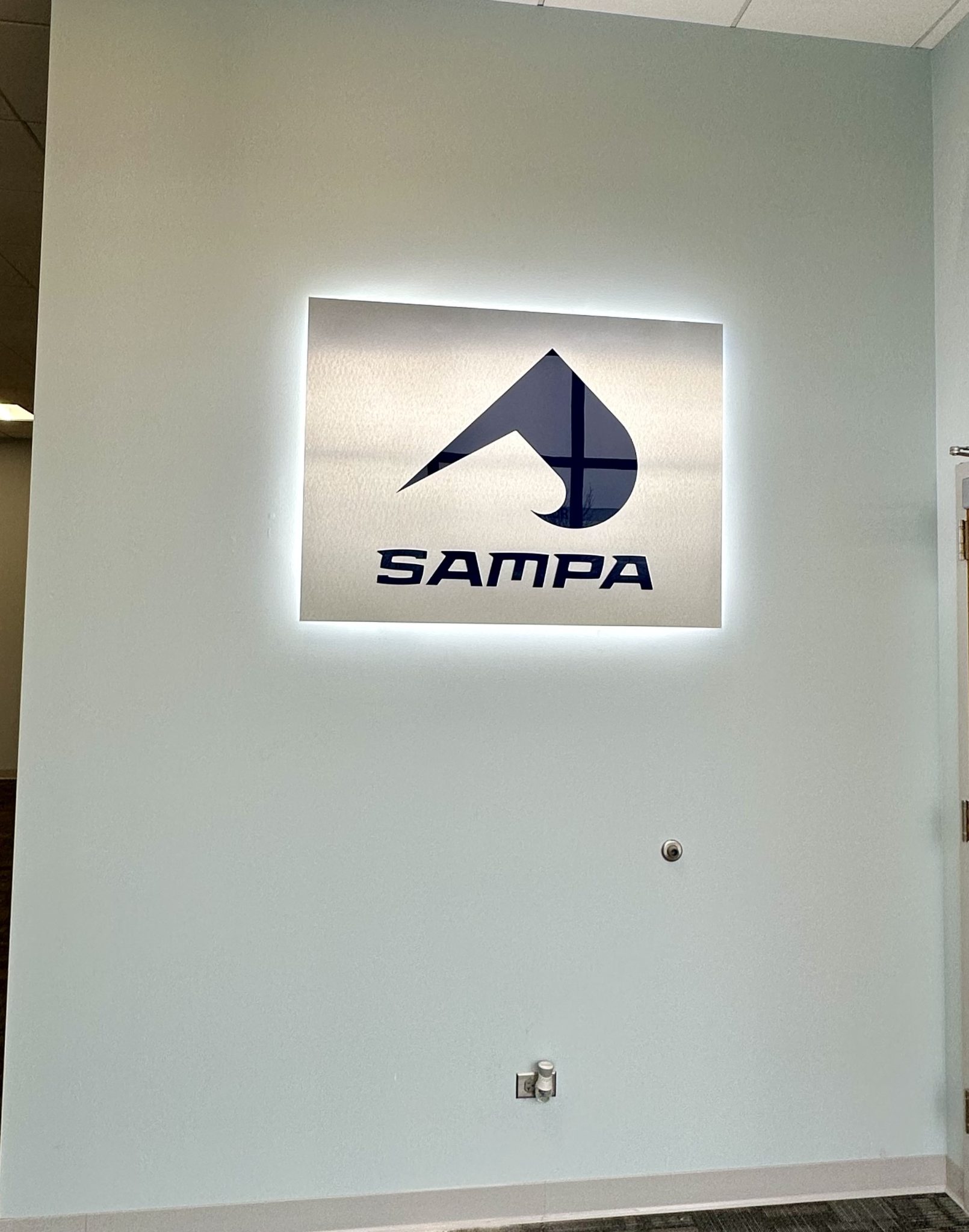 Illuminated acrylic sign displaying the SAMPA logo mounted on a light blue wall.