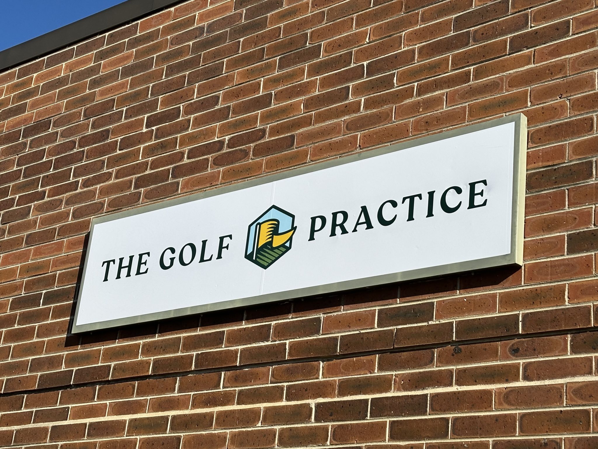 Acrylic exterior sign for The Golf Practice featuring green and gold graphic elements on a brick wall.