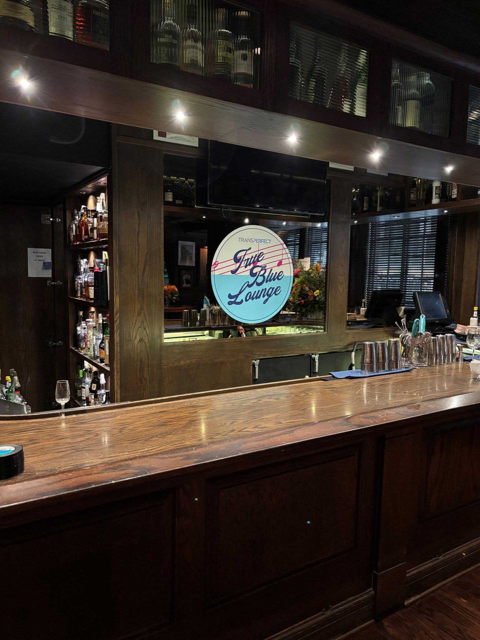 Illuminated circular sign for the True Blue Lounge displayed above a wooden bar, featuring light blue and red typography.