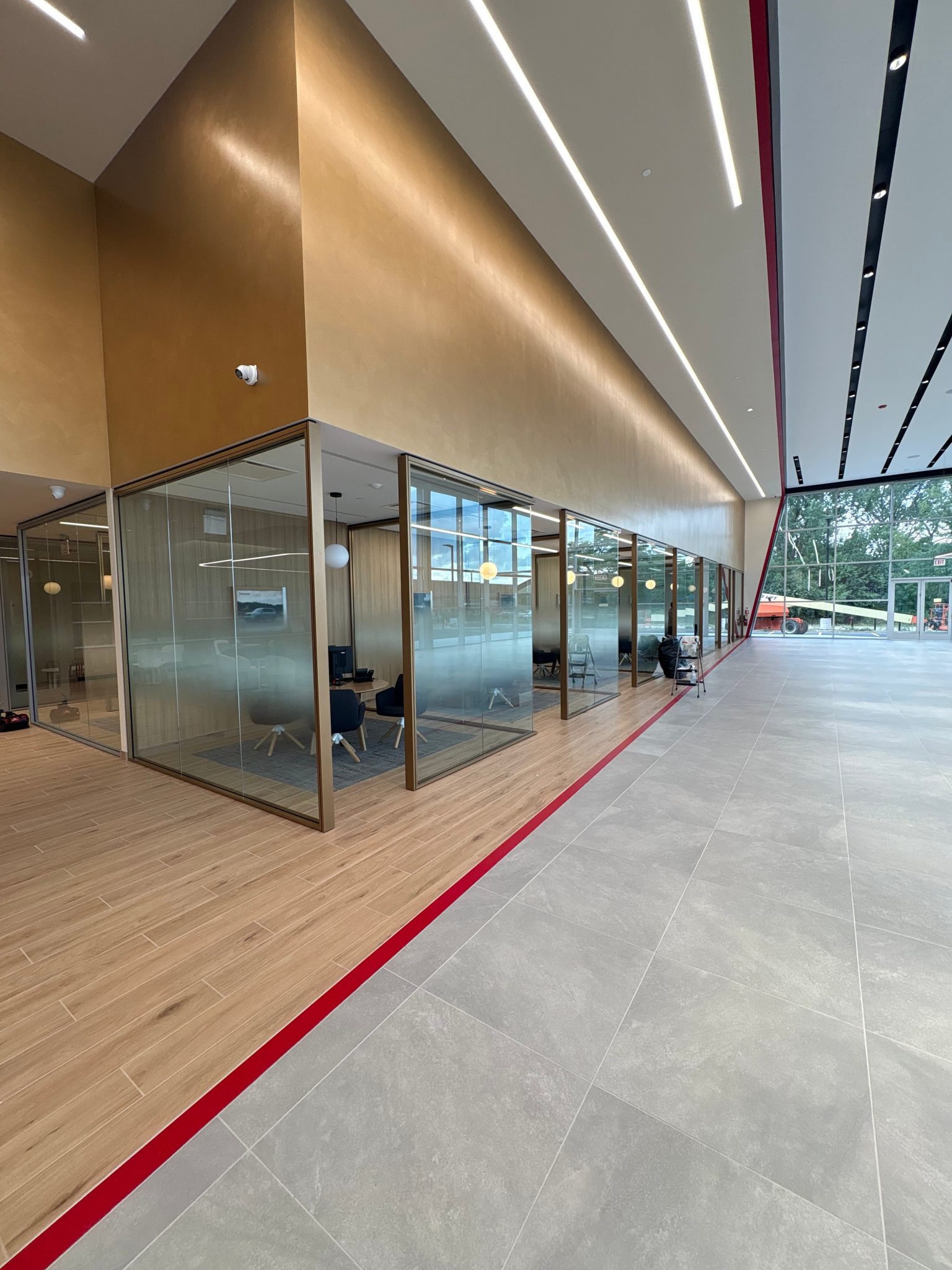 Interior view of an office space featuring glass conference rooms, wooden flooring, and a red accent line along the floor.