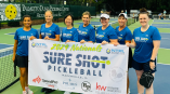 Team of female pickleball players in blue shirts holding a banner for 'Sure Shot Pickleball' at a tournament in Hilton Head, SC.
