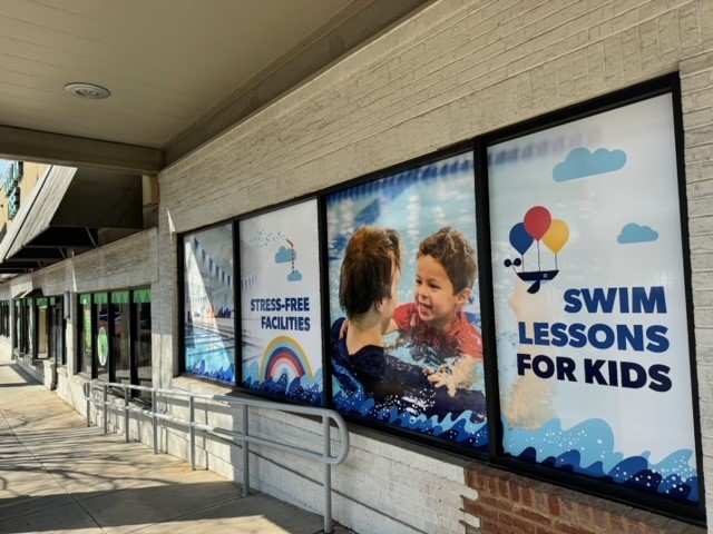 Window graphics promoting swim lessons for kids, featuring cheerful children in water and colorful balloons.