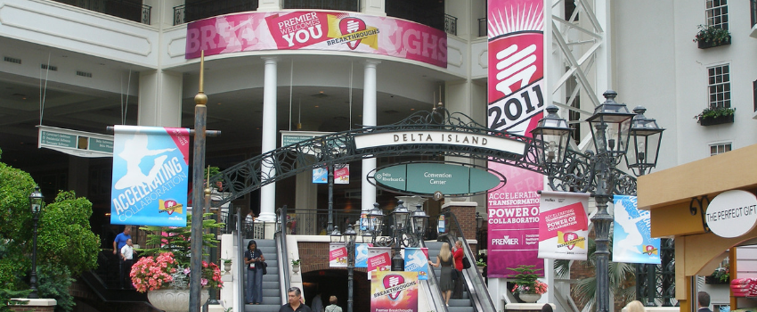 Large pink banner hanging from pillar near elevators outside a convention center.