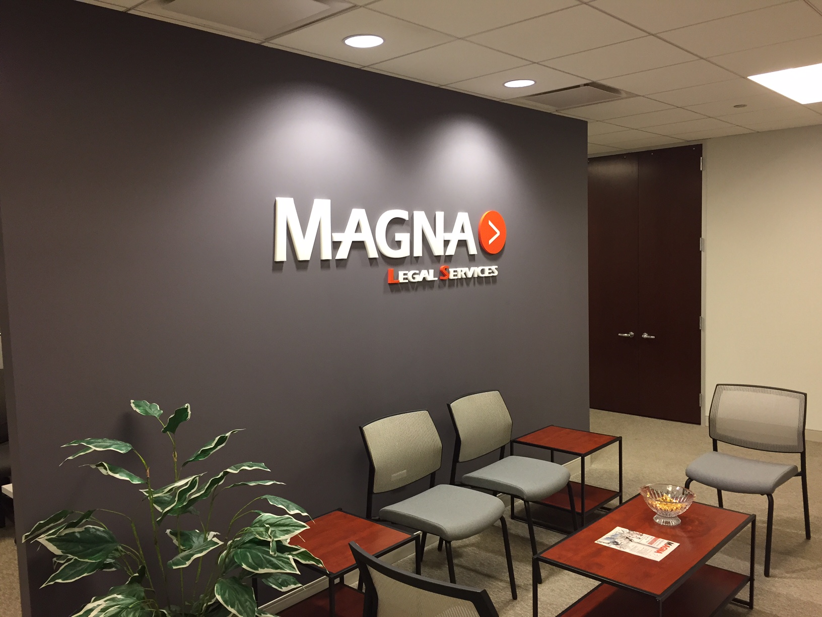 Office waiting room with chairs, a table and a plant in front of a dark wall that says Magna Legal Services in white.